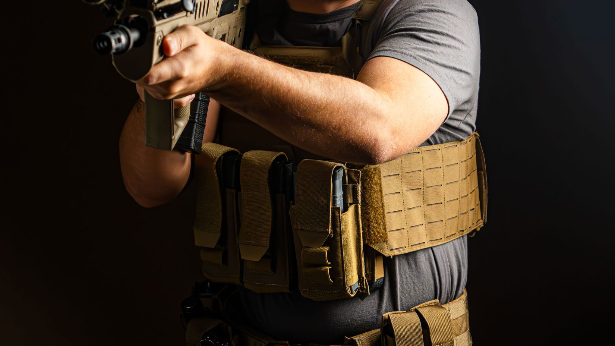 A tactical operator wearing a coyote brown plate carrier and battle belt, equipped with rifle magazine pouches and MOLLE attachments, while aiming a rifle in a combat-ready stance.