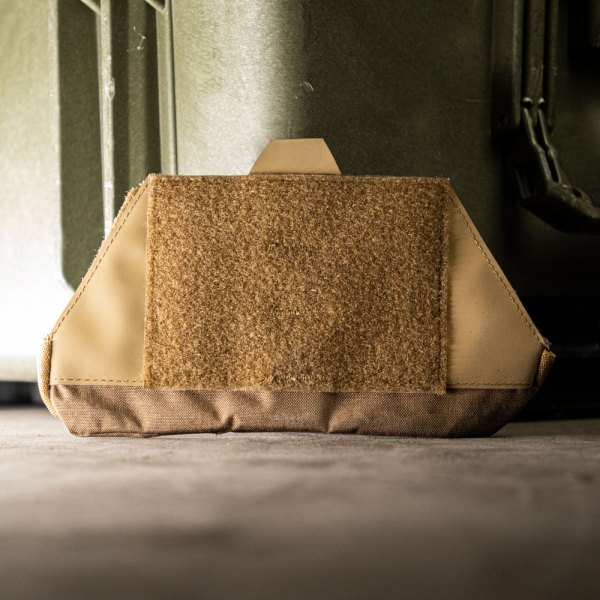 Coyote tactical admin pouch with hook-and-loop panel, designed for mounting on gear or equipment, placed on a rugged surface with a storage case in the background.
