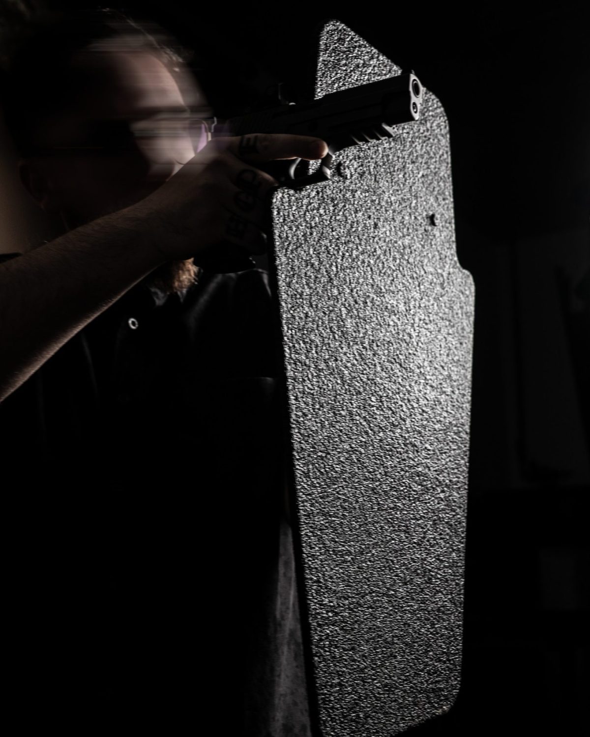 A tactical operator holding a ballistic shield while aiming a handgun, demonstrating personal defense and law enforcement protection in low-light conditions.