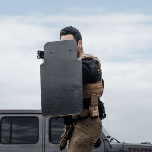 A tactical officer holding a ballistic shield for protection, standing in front of a vehicle in an outdoor environment.